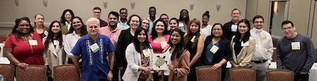 Group photo in a hotel ballroom of 25 attendees of the 2023 DiverseScholar Conference. People are arranged in approximately 3 rows with the two women in front holding a placard of the DiverseScholar green turtle logo. The individuals represent a variety of heights, hair colors, and clothing style with latter mostly business casual.