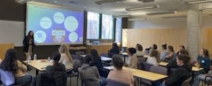 Photo of a WIR event where around 18 women are in a classroom seated in groups of about 4 persons per small table. All are facing a speaker at the front of the room who is standing before a projected slide presentation.